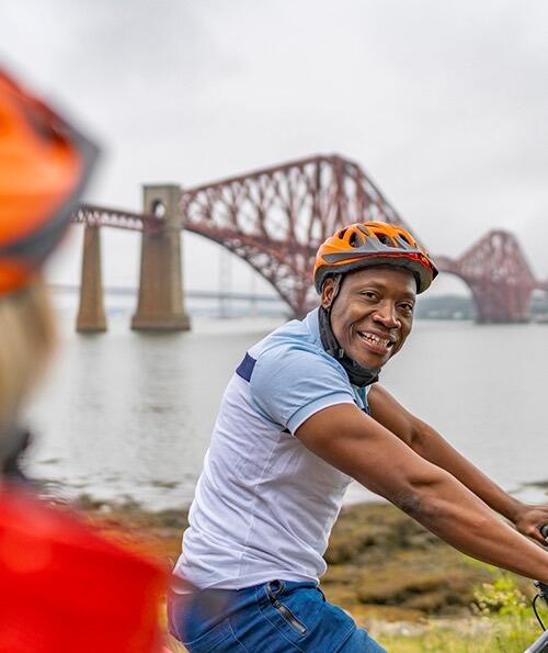 Zwei Radfahrer an der Küste mit der Forth Bridge im Hintergrund.
